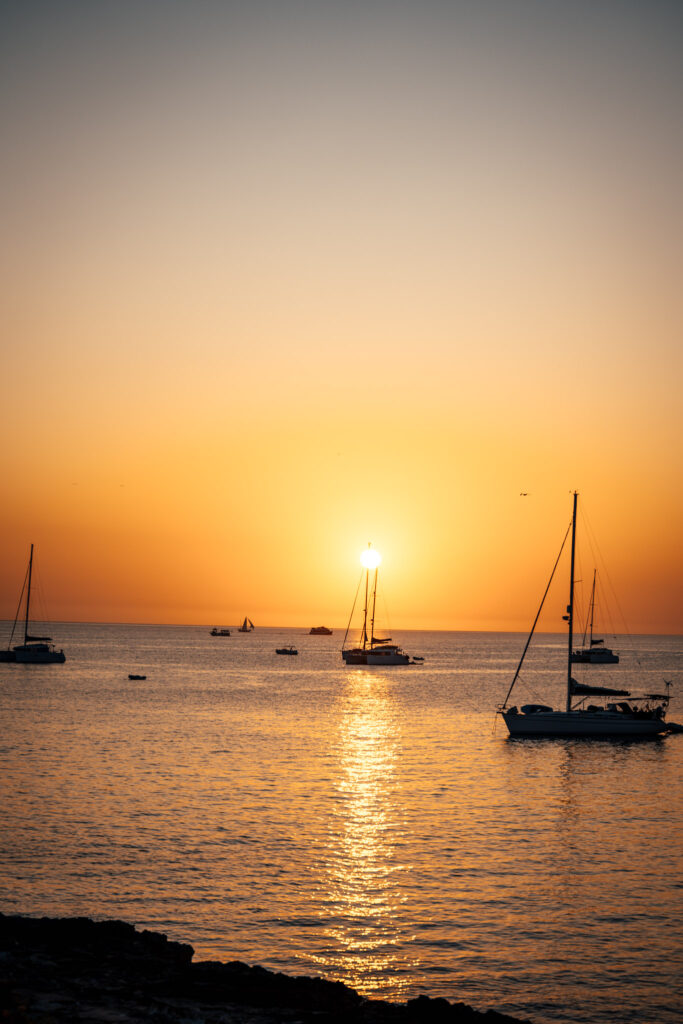 Photo du coucher de soleil, sur la mer, à l’heure dorée. Il y a plusieurs bateaux à voile. Le mat de l’un d’entre eux traverse le soleil.